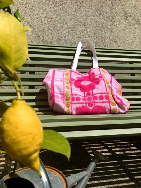 Cabas en tissu éponge motif fleurs, rose, posé sur un banc au soleil à côté d'un citronnier.