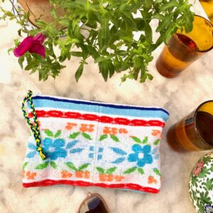 Trousse en tissu éponge motif fleurs, multicolore à dominante bleue, posée sur une table de jardin à coté de verres, de lunettes de soleil et d'une plante fleurie.