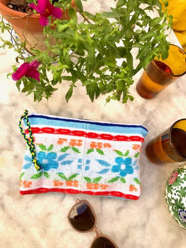 Trousse en tissu éponge motif fleurs, multicolore à dominante bleue, posée sur une table de jardin à coté de verres, de lunettes de soleil et d'une plante fleurie.