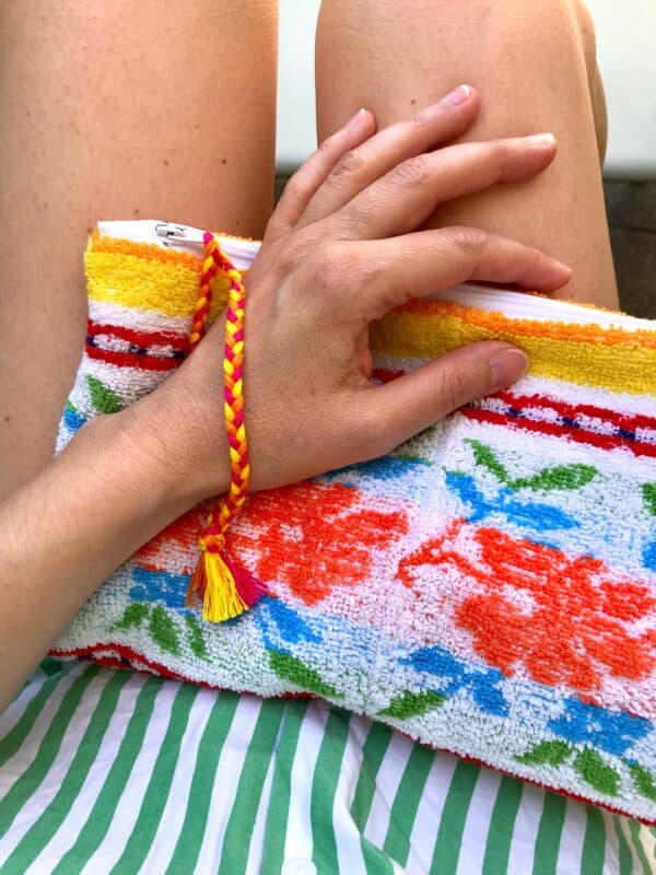 Trousse en tissu éponge motif fleurs, multicolore à dominante jaune, posée sur des jambes nues et passée au poignet grâce à une dragonne tressée tricolore.