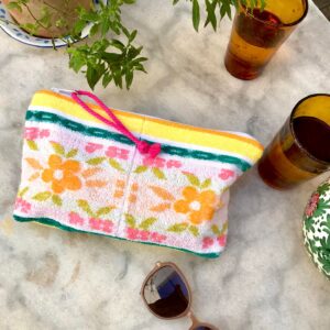 Trousse en tissu éponge motif fleurs, multicolore à dominante jaune et verte, posée sur une table de jardin à coté de verres, de lunettes de soleil et d'une plante fleurie.