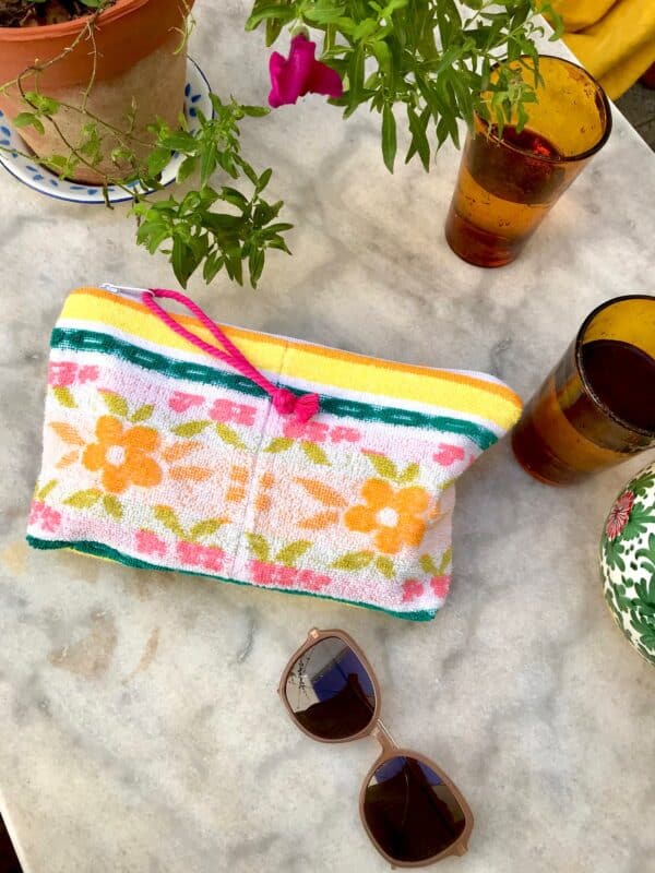 Trousse en tissu éponge motif fleurs, multicolore à dominante jaune et verte, posée sur une table de jardin à coté de verres, de lunettes de soleil et d'une plante fleurie.