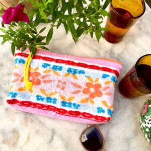 Trousse en tissu éponge motif fleurs, multicolore à dominante rose, posée sur une table de jardin à coté de verres, de lunettes de soleil et d'une plante fleurie.
