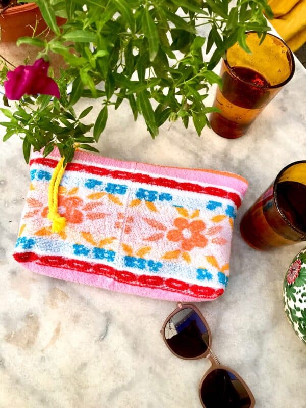 Trousse en tissu éponge motif fleurs, multicolore à dominante rose, posée sur une table de jardin à coté de verres, de lunettes de soleil et d'une plante fleurie.