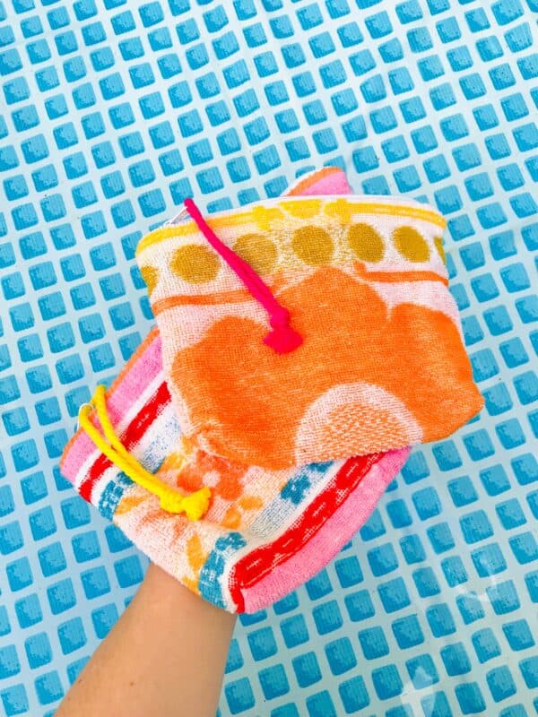 Trousse en tissu éponge motif fleur orange posée sur une autre trousse en éponge et tenues au-dessus d'une piscine à fond bleu.