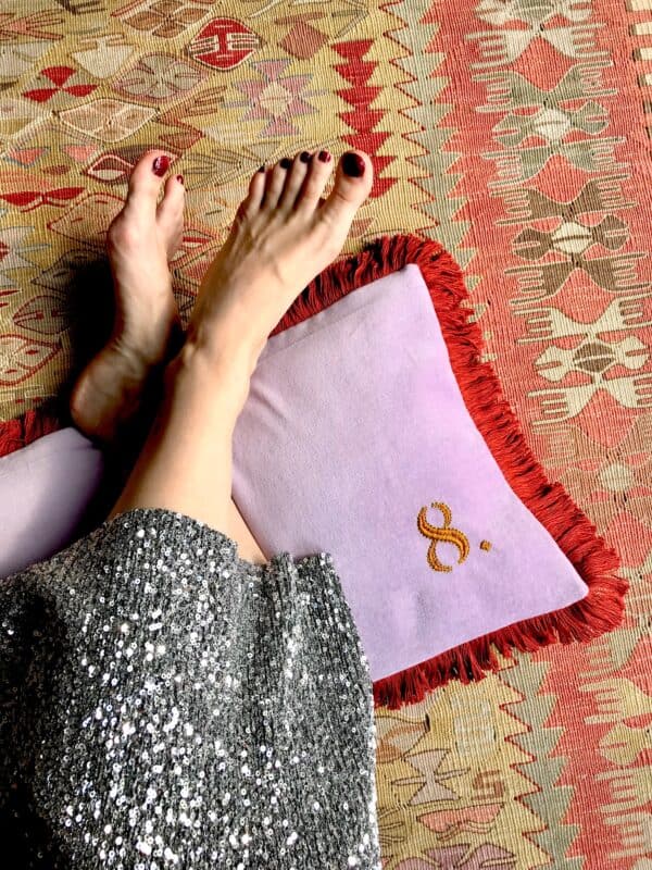 pieds nus croisés sur un coussin en velours lilas, brodé d'un 8 ocre jaune et bordé de franges rouille, sur fond de tapis kilim.