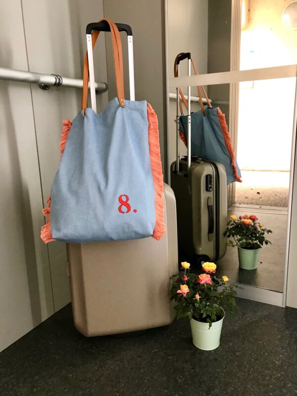 sac cabas bleu délavé, brodé d'un 8 rouge orangé, avec franges vieux roses de chaque côté, suspendu grâce à ses anses en cuir fauve à la poignée d'une valise beige, debout dans un ascenseur.