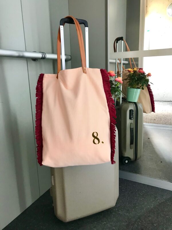sac cabas rose pêche, brodé d'un 8 vert kaki, avec franges bordeaux de chaque côté, suspendu grâce à ses anses en cuir fauve à la poignée d'une valise beige, debout dans un ascenseur.